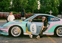 Person sitting in a colorful Porsche car with open door, surrounded by trees and a fence.